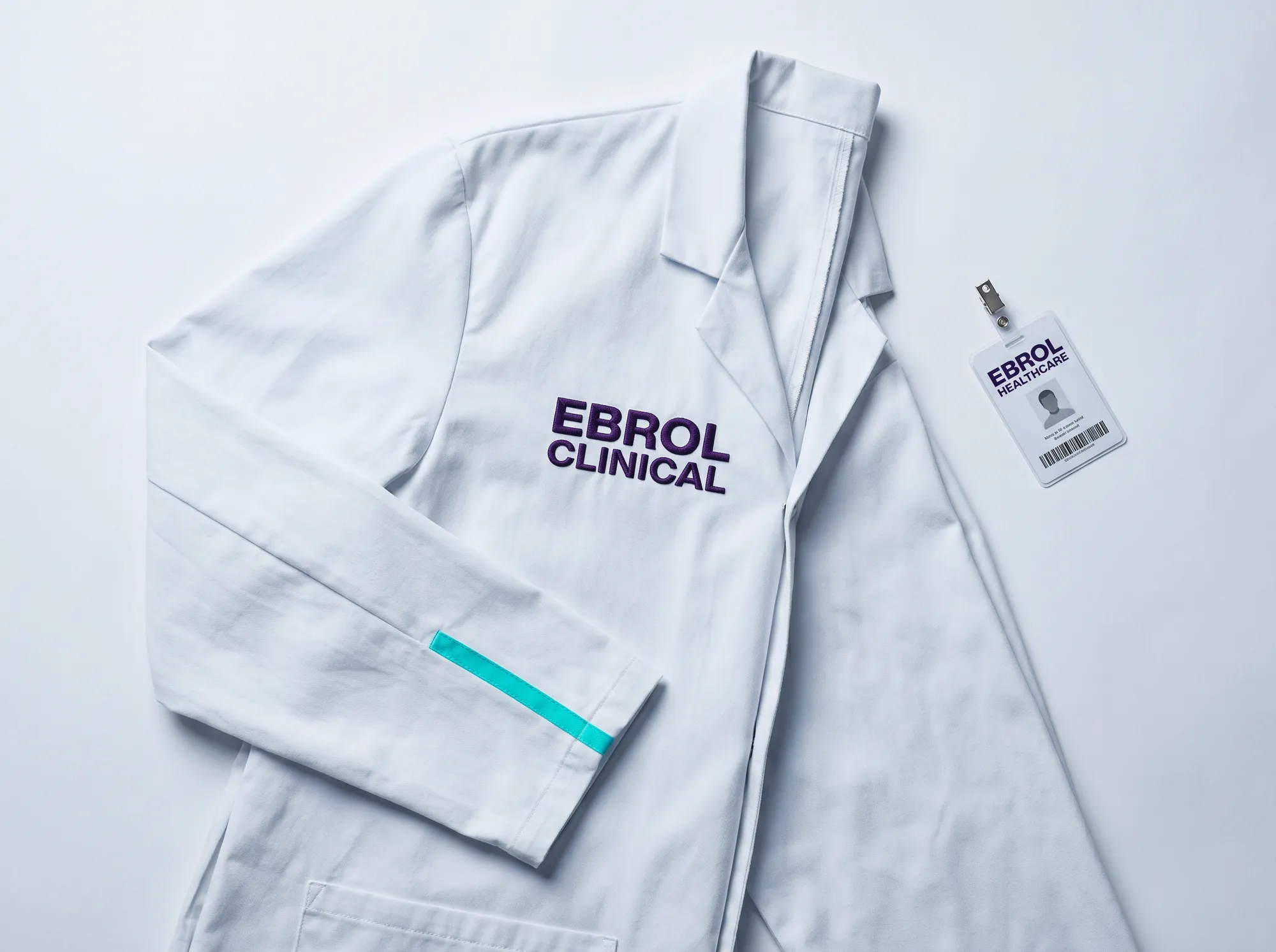 Ebrol Clinical Field Engineer Coat (Apparel) — A working clinical-environment-appropriate field-engineer coat — soft cotton in cool near-white (#FAFAFB) with EBROL CLINICAL embroidered in deep violet (#2A1240) Space Grotesk bold caps above the heart, electric cyan (#00E5C5) accent at the cuff.